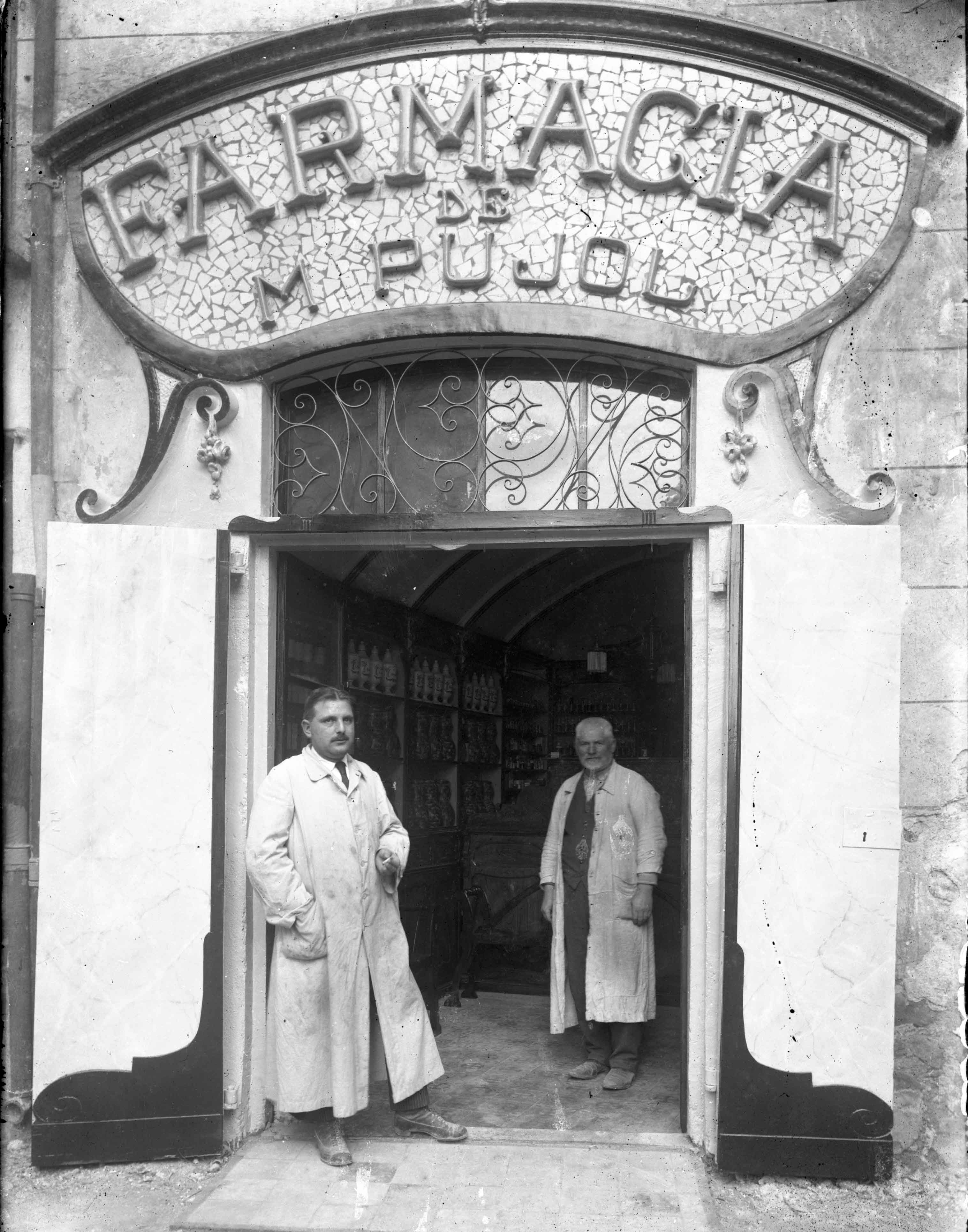 Retrat de cos sencer de dos homes a l'entrada de la farmàcia de Martí Pujol, a Tortellà, 1918 (ACGAX. Fons Sadurní Brunet Pi. Foto: Sadurní Brunet. Codi atribuït 28.1.3)